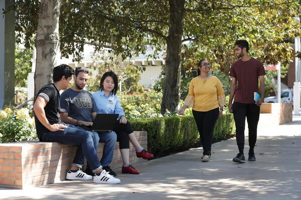 Three students sitting on a wall looking at a computer and two people walking past them