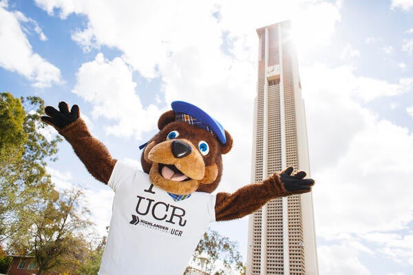 UCR Scotty Bear mascot wearing I Love UCR tshirt with arms raised in front of belltower on UCR campus.