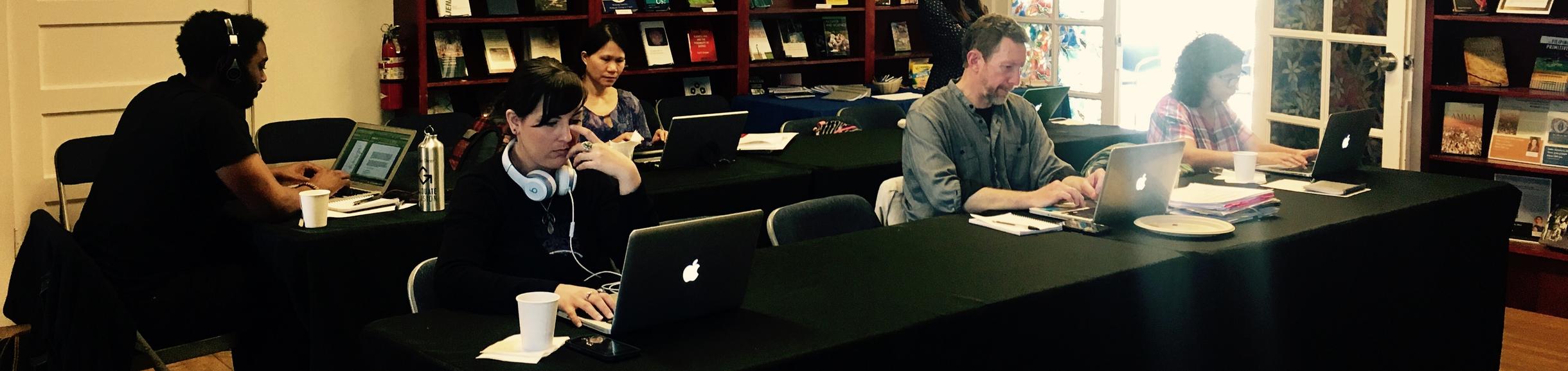 six people in a room with three at a long table in the front on laptops writing